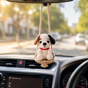 May include: A crocheted puppy ornament hanging from a rearview mirror. The puppy is cream-colored with one brown ear and one black ear, wearing a red collar. It sits on a small, tan crocheted swing suspended by two tan cords with a tassel at the base.