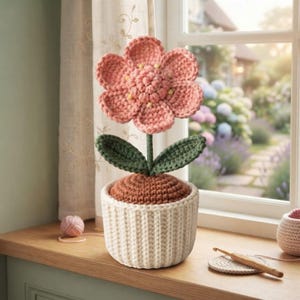 May include: A crocheted flower with pink petals and a textured centre, attached to a green stem with two leaves. The flower is planted in a brown crocheted base inside a ribbed, cream-coloured pot. A wooden crochet hook rests on the surface nearby.