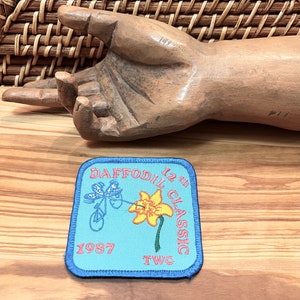 May include: Blue and white embroidered patch with a yellow daffodil and a bicycle with a rider. The text on the patch reads "Daffodil 12th Classic 1987 TWC".