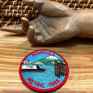 May include: A red, white, and blue embroidered patch with the words "Peninsula Metric 1985" and a depiction of a ferry boat and a mountain.