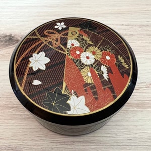 May include: A black round box with a gold and red floral design. The box has a traditional Japanese pattern with flowers, leaves, and a gold ribbon.