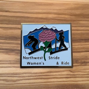 May include: A gold-colored pin with a white background and a blue sky with mountains. The pin features a pink flower with green leaves and two silhouettes of people walking. The text "Northwest Women's Stride & Ride" is printed on the pin.
