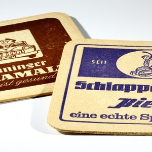 May include: Two vintage cardboard coasters. The first coaster is brown with a white image of a cartoon man driving a car and the text "Henninger KARAMAL ...das ist gesund". The second coaster is beige with a blue image of a cartoon man holding a beer mug and the text "SEIT 16 31 Schlappefeppel Bier eine echte Spezialität!"