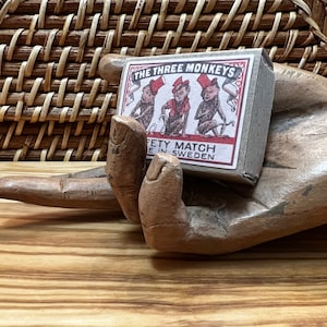 May include: A vintage matchbox with the text "THE THREE MONKEYS" and illustrations of monkeys in hats and clothing. The matchbox is held by a carved wooden hand. The background includes a woven basket and a wooden surface.
