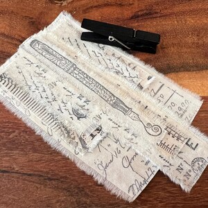 May include: A stack of fabric scraps with a frayed edge, featuring a vintage design with a pen and ink illustration. The fabric is a light beige color with black ink.
