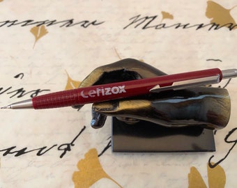 May include: A burgundy mechanical pencil with the word "Cerizox" printed on the side, resting on a black hand-shaped pen holder. The pencil has a silver tip and clip. The background is a patterned paper.