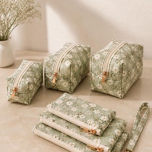 Könnte beinhalten: Ein Set aus salbeigrünen Stofftaschen mit weißem Gänseblümchenmuster. Das Set enthält drei rechteckige, boxförmige Taschen mit cremefarbenen Reißverschlüssen sowie drei flache, rechteckige Taschen, von denen eine mit einer Handschlaufe versehen ist.