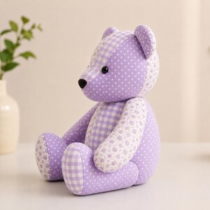 Puede incluir: Un oso de peluche hecho de tela color lavanda con una mezcla de patrones que incluyen lunares blancos, un estampado de cuadros vichy cl&aacute;sico y un peque&ntilde;o dise&ntilde;o floral. El oso tiene dos peque&ntilde;os ojos de pl&aacute;stico negro y una nariz negra bordada.