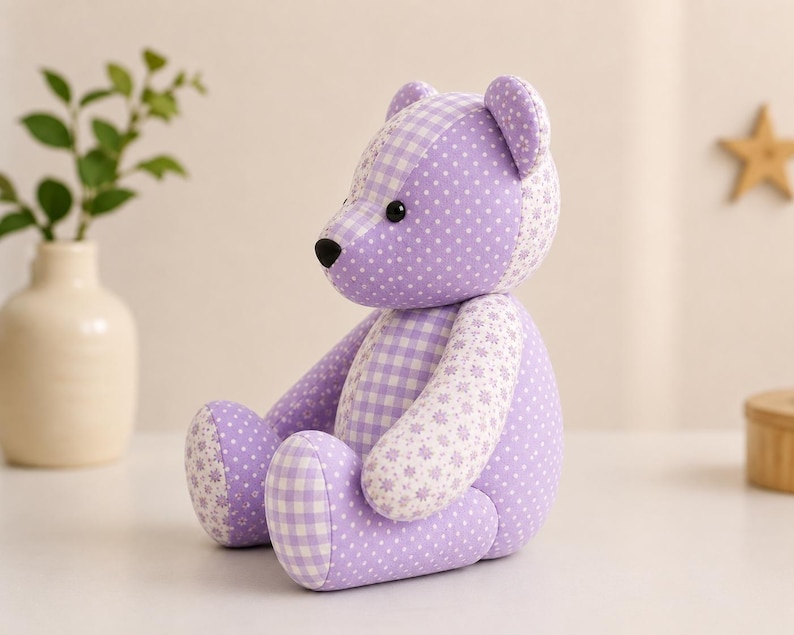 Puede incluir: Un oso de peluche hecho de tela color lavanda con una mezcla de patrones que incluyen lunares blancos, un estampado de cuadros vichy cl&aacute;sico y un peque&ntilde;o dise&ntilde;o floral. El oso tiene dos peque&ntilde;os ojos de pl&aacute;stico negro y una nariz negra bordada.