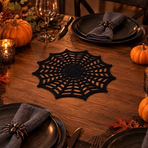 May include: A black crocheted spider web placemat with a circular center and radiating web patterns, positioned on a wooden table set with dark plates, grey napkins, spider napkin rings, orange pumpkins, and lit black taper candles.