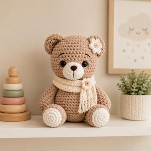 May include: A light brown crocheted teddy bear with a cream-coloured scarf, a small white flower on its ear, and white circular patches on its feet. The bear has dark eyes, a brown stitched nose, and pink blush accents on its cheeks.