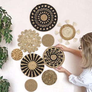 May include: A collection of nine round woven wall baskets and decorative discs in natural tan and black fibers. The set includes various textures, including a sunburst design with cream tassels, spiral patterns, and solid braided circular mats.