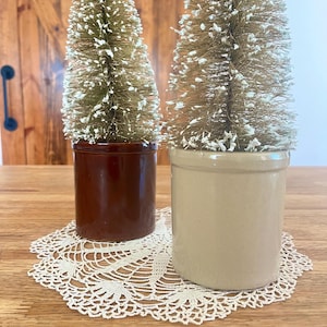 May include: Two bottle brush trees with white-tipped needles are set in cylindrical ceramic crocks. One crock is dark brown and the other is light beige. The trees are placed on a white crocheted doily with a detailed floral pattern.