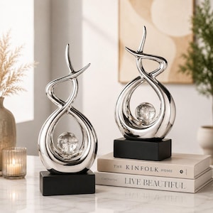 May include: Two silver-toned abstract sculptures featuring fluid, looping forms that enclose a clear glass sphere with internal bubbles. Each sculpture is mounted on a solid black rectangular base, creating a balanced and reflective display piece.