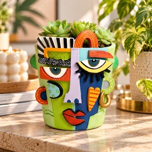 May include: A ceramic planter shaped like a human face with a split color design. The left side is light green with black and white patterns, while the right side is dark blue. It features a light purple nose, red lips, and a succulent plant.