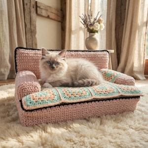 May include: A light pink crocheted pet sofa featuring a seat cushion made of square granny motifs in shades of mint green and peach. The sofa has a solid pink backrest and armrests with a black trim along the edges, providing a soft resting area.