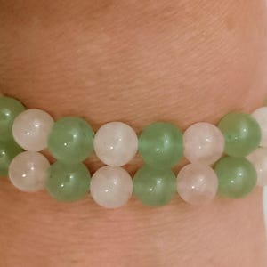 Puede incluir: Una pulsera de doble hilo compuesta por cuentas redondas y lisas que alternan entre rosa pálido y verde salvia translúcido. Las cuentas están ensartadas en un patrón repetitivo, creando dos filas paralelas de piedras pulidas alrededor.