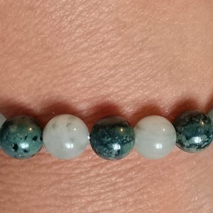 Puede incluir: Una pulsera de cuentas con esferas alternas en tonos verde bosque oscuro con motas negras y cuentas lisas y transl&uacute;cidas de color blanco p&aacute;lido o verde menta claro. Las cuentas est&aacute;n ensartadas en un patr&oacute;n repetitivo sobre un cord&oacute;n el&aacute;stico.