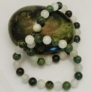 Puede incluir: Una pulsera de cuentas con cuentas redondas pulidas que alternan tonos de verde bosque, oliva oscuro y blanco transl&uacute;cido. Las cuentas est&aacute;n ensartadas en un patr&oacute;n circular, mostrando un acabado suave y brillante en cada piedra individual.