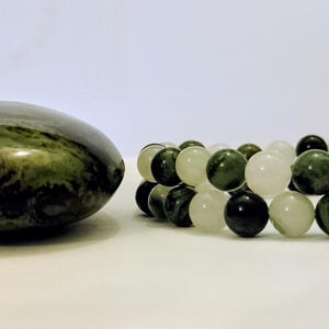 Puede incluir: Una piedra pulida de color verde oscuro con patrones en espiral se encuentra junto a una pulsera de dos hilos. La pulsera est&aacute; compuesta por cuentas redondas alternas en tonos de blanco transl&uacute;cido, verde bosque oscuro y piedra verde moteada.