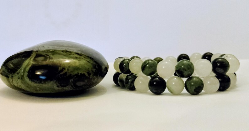 Puede incluir: Una piedra pulida de color verde oscuro con patrones en espiral se encuentra junto a una pulsera de dos hilos. La pulsera est&aacute; compuesta por cuentas redondas alternas en tonos de blanco transl&uacute;cido, verde bosque oscuro y piedra verde moteada.