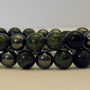 Puede incluir: Una pulsera de doble hilo compuesta por cuentas redondas alternas en verde bosque oscuro con texturas moteadas y esferas metálicas de color gris carbón pulido. Las cuentas están ensartadas en un patrón repetitivo formando dos filas paralelas.