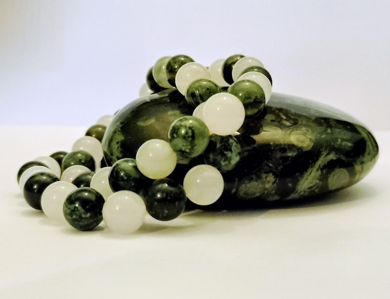 Puede incluir: Una pulsera de cuentas con esferas lisas que alternan entre un verde bosque profundo con patrones oscuros y un blanco roto transl&uacute;cido. La pulsera est&aacute; dispuesta en forma circular y descansa sobre una piedra grande de color verde oscuro.