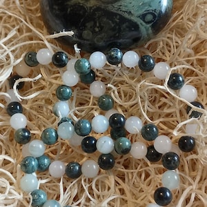 Puede incluir: Una piedra pulida en forma de orbe, de color verde oscuro con patrones circulares, se sit&uacute;a sobre dos pulseras de cuentas circulares. Las pulseras est&aacute;n compuestas por una alternancia de cuentas blancas lisas y transl&uacute;cidas.