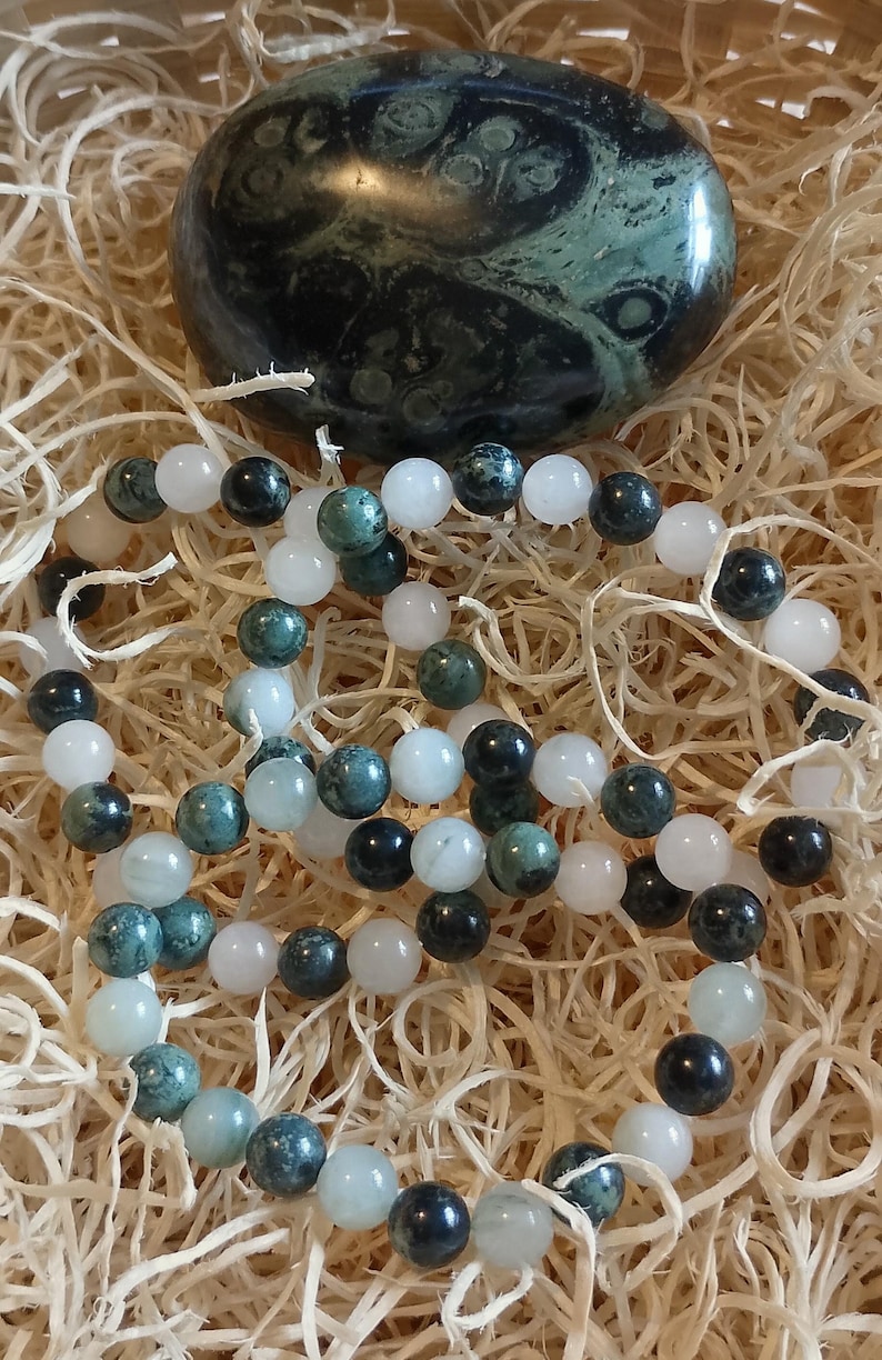 Puede incluir: Una piedra pulida en forma de orbe, de color verde oscuro con patrones circulares, se sit&uacute;a sobre dos pulseras de cuentas circulares. Las pulseras est&aacute;n compuestas por una alternancia de cuentas blancas lisas y transl&uacute;cidas.