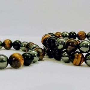 Puede incluir: Dos pulseras elásticas compuestas por cuentas esféricas alternas en negro pulido, gris metálico y piedra ojo de tigre marrón dorado. Las cuentas están dispuestas en un patrón repetitivo, creando una textura suave y uniforme en las bandas.