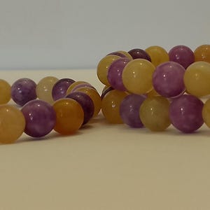 Puede incluir: Dos pulseras elásticas hechas de cuentas redondas y pulidas que alternan tonos amarillo dorado y morado intenso. Las cuentas están ensartadas en un cordón elástico, creando una forma circular. Las pulseras tienen un acabado liso y brillante.