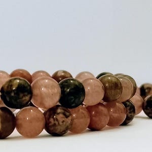 Puede incluir: Dos pulseras de cuentas compuestas por piedras esféricas lisas en tonos variados de rosa empolvado, marrón apagado y carbón oscuro. Las cuentas están ensartadas en un patrón circular continuo, mostrando variaciones de color naturales.