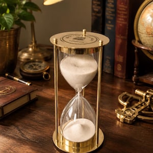 May include: A brass hourglass with clear glass bulbs filled with white sand. The top circular brass plate features an engraved ship illustration and the text "BELL FOR SERVICE" and "LONDON" in a circular pattern around the central image.