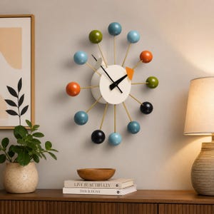 Può includere: Un orologio da parete con un quadrante circolare bianco centrale, lancette nere e un piccolo accento triangolare arancione. Dodici sottili aste metalliche color oro si estendono verso l'esterno, ciascuna sormontata da una sfera blu, verde, arancione o nera.