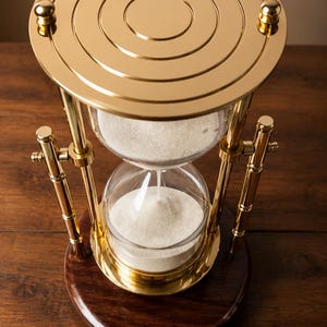 Peut inclure: Un sablier doté d'une structure en métal doré poli avec des détails circulaires concentriques sur la plaque supérieure. Les bulbes en verre transparent contiennent du sable blanc fin, le tout reposant sur une base en bois brun foncé.