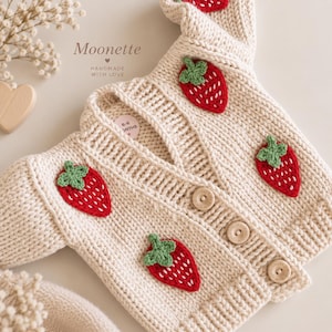 May include: A cream-colored knit cardigan featuring a V-neckline, long sleeves, and three light wooden buttons. The garment is decorated with several red and green crocheted strawberry appliques. A small circular tag indicates 6-6 MONTHS.