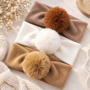 Può includere: Tre fasce per capelli in maglia a coste, ciascuna dotata di un grande e soffice pon-pon in pelliccia sintetica. Le fasce sono di colore marrone scuro, bianco crema e marrone chiaro, disposte in pila verticale con i pon-pon centrati.