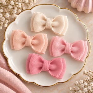 Peut inclure: Quatre nœuds pour cheveux en tissu disposés sur une assiette blanche festonnée. Les nœuds présentent une texture côtelée et se déclinent dans un dégradé de couleurs crème, pêche clair, rose pâle et rose soutenu, noués par un centre.