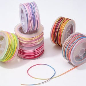 May include: Five clear plastic spools wound with multi-colored cord. One spool features pink, purple, and blue tones. Another has yellow and green. A stack of three spools displays pink, red, and yellow hues. A loose strand of rainbow cord lies below.