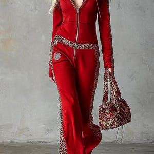 Könnte beinhalten: Ein roter Jumpsuit mit Reißverschluss, Kapuze, langen Ärmeln und weit geschnittener Hose, akzentuiert durch seitliche Einsätze und Besätze mit Leopardenmuster. Das Kleidungsstück hat einen silbernen Frontreißverschluss und ein silbernes Emblem.