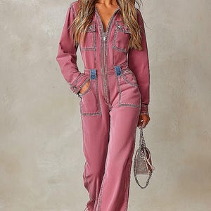 Könnte beinhalten: Ein altrosa Jeans-Jumpsuit mit einer Kapuze mit dunkelblauem Jeansfutter, einem durchgehenden Frontreißverschluss, langen Ärmeln und mehreren aufgesetzten Taschen. Das Kleidungsstück verfügt über kontrastierende blaue Jeans-Akzente an der Taille.