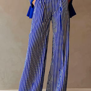 May include: High-waisted wide-leg trousers in royal blue with vertical white pinstripes, featuring a gold-toned circular buckle belt and a matching royal blue blazer with gold buttons. The trousers have a structured waistband and a flowing silhouette.