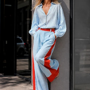 May include: A light blue and white vertical striped long-sleeve button-down shirt with a collar, paired with wide-leg light blue trousers featuring bold red side stripes and a red hem. The outfit includes red-tinted sunglasses and silver earrings.