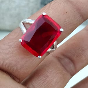 May include: A silver ring featuring a large, rectangular, faceted red gemstone held in place by four metal prongs. The band of the ring has a split design that connects to the setting, showcasing a polished metallic finish and a vibrant red stone.
