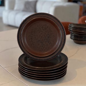 Może przedstawiać: Stos okrągłych ceramicznych talerzy o głębokim brązowym środku z nakrapianą fakturą i ciemniejszym, jednolitym brązowym brzegiem. Talerze mają gładkie, szkliwione wykończenie i lekko uniesiony brzeg, a jeden talerz stoi pionowo przed stosem.