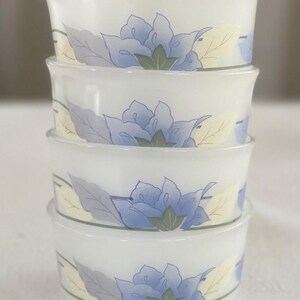 May include: A stack of four white milk glass bowls featuring a repeating floral pattern. Each bowl displays a central blue flower with green leaves, flanked by pale yellow and grey foliage, all connected by a thin horizontal line encircling the rim.