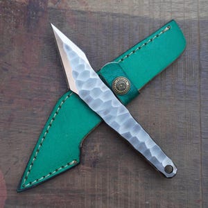 May include: A fixed-blade knife with a silver-toned, hammered-texture handle and a pointed blade. The knife is paired with a bright green sheath featuring visible stitching along the edges and a circular brass-coloured metal snap button closure.