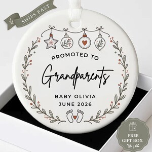 May include: A round white ceramic ornament featuring a black and red botanical wreath design. The text reads PROMOTED TO Grandparents, BABY OLIVIA, and JUNE 2026. Above the text are four hanging ornaments, and below are two baby footprints.