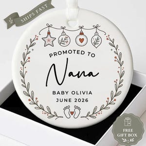 May include: A round white ceramic ornament featuring a black text design that reads PROMOTED TO Nana, BABY OLIVIA, and JUNE 2026. The ornament is decorated with a botanical wreath, hanging holiday baubles, and two baby footprints with a heart.