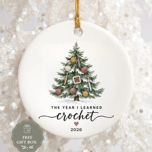 May include: A round white ceramic ornament featuring a watercolor illustration of a Christmas tree decorated with yarn balls and crochet hooks. The text reads THE YEAR I LEARNED crochet with a small pink heart and the year 2026 printed below.